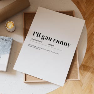 May include: A white print with the phrase "I'll gan canny" in black, with the phonetic pronunciation and definition. The print is in a wooden frame, with a cup of coffee and a book visible.
