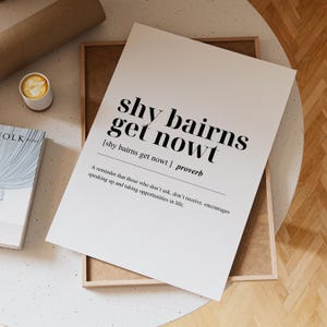 以下が含まれることがあります： 「shy bairns get nowt」の黒い文字が書かれた白いプリント。定義とリマインダーが添えられています。木製のフレームに入っており、コーヒーカップと本が近くにあります。