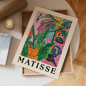 May include: A framed art print with a vibrant abstract floral design in pink, green, orange, and blue. The print is labelled "MATISSE" in bold black letters. A cup of coffee and a book are visible in the background.