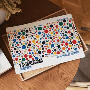 Könnte beinhalten: Ein Druck mit einem bunten Punktmuster in Rot, Blau, Gelb, Grün und Schwarz. Der Druck enthält den Text "YAYOI KUSAMA" und "Japan 1976". Das Kunstwerk wird auf einem Holztablett präsentiert.
