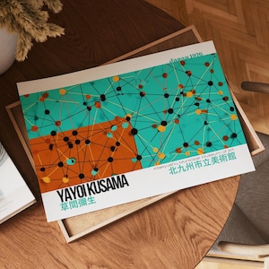 May include: A print featuring a colorful abstract design with teal, orange, black, and yellow elements. The print includes the text "YAYOI KUSAMA" and Japanese characters, along with "Kitakyushu Municipal Museum of Art" and "Japan 1976". The artwork is displayed on a wooden surface.