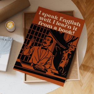 Peut inclure: Une affiche orange avec le texte "I speak English well. I learn it from a book !" présente une illustration noire et orange d'un homme en costume et d'un élan. L'affiche est sur un plateau en bois, avec une tasse de café et un livre à proximité.