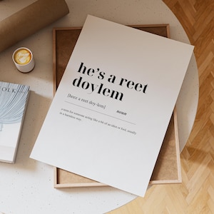 Peut inclure: Impression blanche avec le texte "he's a reet doylem" en noir, avec une définition en dessous. L'impression est dans un cadre en bois, avec une tasse de café et un livre visibles.