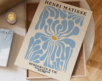 Puede incluir: Una impresión artística enmarcada con un fondo crema, que presenta una flor azul estilizada con un centro amarillo. La impresión incluye el texto "HENRI MATISSE" y "BERGGRUEN & CIE" con una dirección. La impresión se muestra sobre una superficie de color claro.