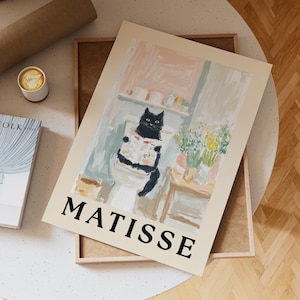 Könnte beinhalten: Ein Druck mit einer schwarzen Katze, die eine Zeitung auf einer Toilette liest. Das Kunstwerk befindet sich in einem Badezimmer mit einer hellen Farbpalette. Das Wort "MATISSE" ist unten in fetten schwarzen Buchstaben gedruckt.