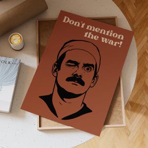 Könnte beinhalten: Ein rostfarbenes Poster mit dem Text "Don't mention the war!" und einer Schwarz-Weiß-Illustration eines Mannes, der zwinkert. Das Poster befindet sich in einem Holzrahmen und liegt auf einem weißen Tisch. Eine Kaffeetasse und ein Buch sind ebenfalls zu sehen.