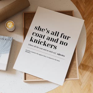 Puede incluir: Impresión en blanco con el texto "she's all fur coat and no knickers" en negro, con una definición debajo. La impresión está en un marco de madera, con una taza de café y un libro cerca.