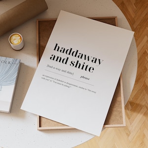 Puede incluir: Impresión en blanco con la frase "haddaway and shite" en negro, junto con su pronunciación y definición. La impresión está en un marco de madera, con una taza de café y un libro cerca.
