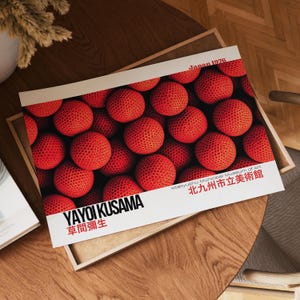 Könnte beinhalten: Ein Druck mit einer Nahaufnahme vieler roter Kugeln, möglicherweise Erdbeeren, mit einer strukturierten Oberfläche. Der Druck enthält den Text "YAYOI KUSAMA" und japanischen Text sowie "Japan 1976" und "Kitakyushu Municipal Museum of Art".