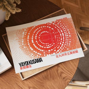 May include: A print featuring a vibrant, abstract design in shades of red and white, with the text "YAYOI KUSAMA" and Japanese characters. The print is labeled "Japan 1976" and "Kitakyushu Municipal Museum of Art". It rests on a wooden tray.