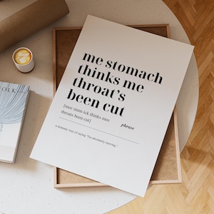 Könnte beinhalten: Ein weißer Druck mit dem schwarzen Text "me stomach thinks me throat's been cut". Der Druck befindet sich in einem Holzrahmen, mit einer Kaffeetasse und einem Buch im Hintergrund. Der Druck ist ein humorvolles Wandkunstwerk.