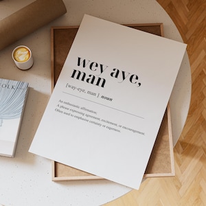 Può includere: Una stampa bianca con la frase "wey aye, man" in testo nero, con una definizione. La stampa è in una cornice di legno, con una tazza di caffè e un libro visibili.