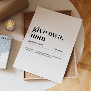 May include: A white print with the phrase "give owa, man" in a bold, black font. The print includes the definition of the phrase and is displayed on a wooden frame. A cup of coffee and a book are also visible.