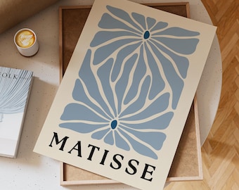 Puede incluir: Una impresión artística enmarcada con un diseño floral azul claro sobre un fondo crema. La impresión tiene la palabra "MATISSE" en negro en la parte inferior. La impresión descansa sobre un marco de madera, con una taza de café y un libro cerca.