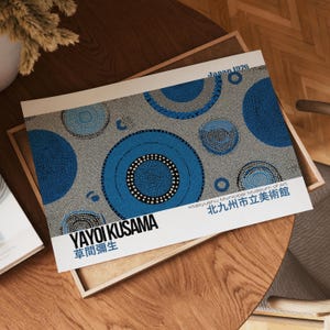 May include: A print featuring Yayoi Kusama's artwork from 1976, with blue and white dotted circles on a gray background. The print includes Japanese text and is displayed on a wooden tray.