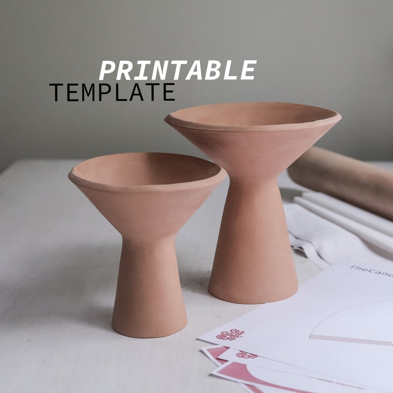 May include: Two terracotta-colored ceramic candle holders, one larger than the other, with a flared top and a wide base. The candle holders are on a white surface.