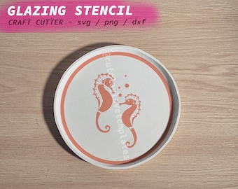 Könnte beinhalten: Ein weißer Keramikteller mit einem korallenorangen Rand und zwei Seepferdchen-Silhouetten in der Mitte. Der Teller liegt auf einer Holzoberfläche.  "Glazing Stencil" und "Craft Cutter ~ svg / png / dxf" sind in rosa Schrift über dem Teller geschrieben.