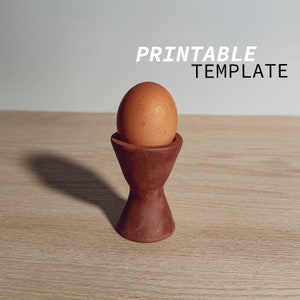 May include: A brown egg in a wooden egg cup on a light brown wooden surface. The text "PRINTABLE TEMPLATE" is visible above the egg.