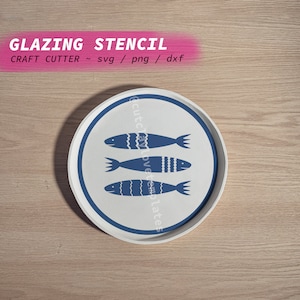 May include: A white ceramic plate with a blue border and three blue fish designs. The plate is on a light brown wooden surface. The text "Glazing Stencil" is at the top of the image. The text "Craft Cutter ~ svg / png / dxf" is below the first line of text.