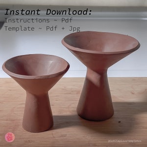 May include: Two terracotta-colored ceramic pedestals with a wide, shallow bowl on top. The pedestals are different sizes, with the larger one in the background and the smaller one in the foreground. The pedestals are on a wooden surface.