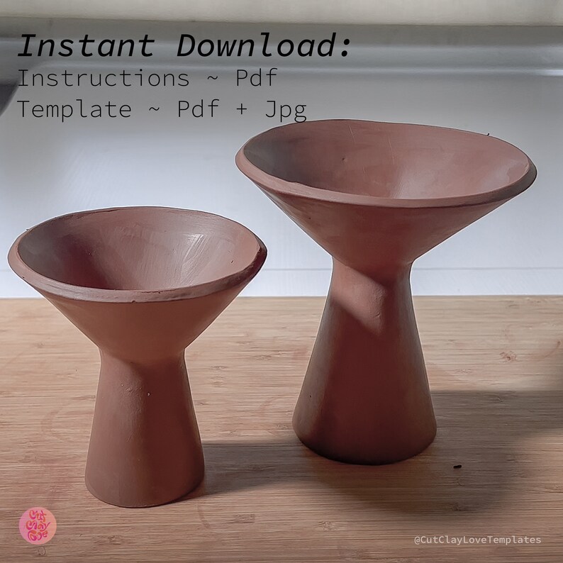 May include: Two terracotta-colored ceramic pedestals with a wide, shallow bowl on top. The pedestals are different sizes, with the larger one in the background and the smaller one in the foreground. The pedestals are on a wooden surface.