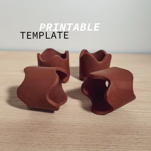 May include: Four terracotta-colored, handmade ceramic holders with a wavy design. The holders are arranged on a light-colored wooden surface. The text "PRINTABLE TEMPLATE" is displayed in white above the holders.