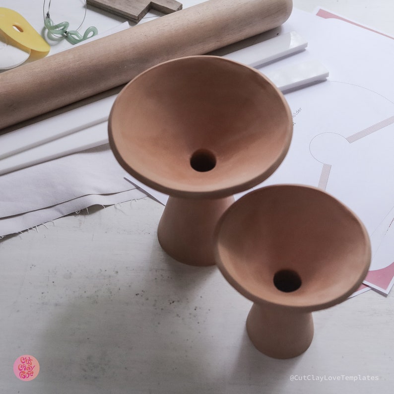 May include: Two terracotta-colored ceramic vases with a wide, flared opening and a narrow base. The vases are sitting on a white surface.