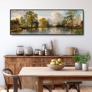 K&ouml;nnte beinhalten: Ein Panorama-Landschaftsgem&auml;lde, das einen See mit B&auml;umen und einer wei&szlig;en Kirche in der Ferne zeigt. Das Gem&auml;lde ist in Schwarz gerahmt und h&auml;ngt an einer wei&szlig;en Wand &uuml;ber einem Holzschrank und einem Tisch.
