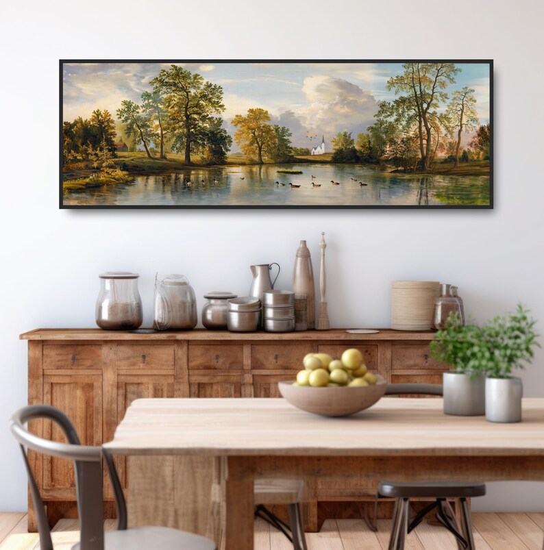 K&ouml;nnte beinhalten: Ein Panorama-Landschaftsgem&auml;lde, das einen See mit B&auml;umen und einer wei&szlig;en Kirche in der Ferne zeigt. Das Gem&auml;lde ist in Schwarz gerahmt und h&auml;ngt an einer wei&szlig;en Wand &uuml;ber einem Holzschrank und einem Tisch.