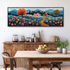 May include: A long, horizontal painting of a whimsical landscape with a pink house, a winding path, and a field of colorful flowers. The painting is framed in black and hangs on a white wall above a wooden table and chairs.