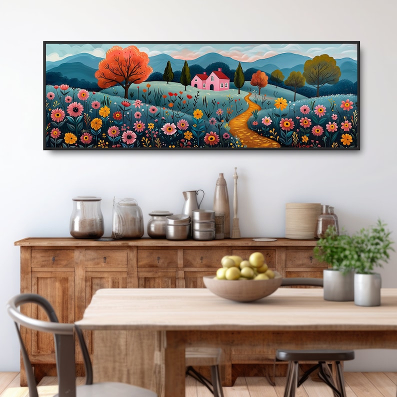 May include: A long, horizontal painting of a whimsical landscape with a pink house, a winding path, and a field of colorful flowers. The painting is framed in black and hangs on a white wall above a wooden table and chairs.