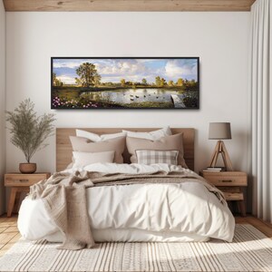 May include: A panoramic landscape painting of a lake with ducks swimming in the water. The painting is framed in black and hangs above a bed with white bedding and a brown throw blanket.