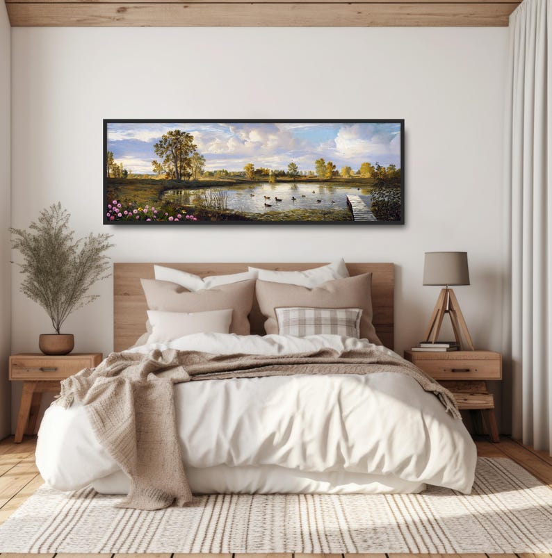 May include: A panoramic landscape painting of a lake with ducks swimming in the water. The painting is framed in black and hangs above a bed with white bedding and a brown throw blanket.