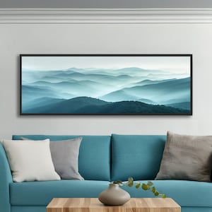Könnte beinhalten: Ein panoramisches Wandkunstbild mit einer Landschaft aus geschichteten Gebirgszügen in Blau- und Petroltönen, die in einem nebligen Horizont verblassen. Das Kunstwerk ist in einem dünnen schwarzen Rahmen gefasst und hängt über einem Sofa.