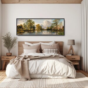 K&ouml;nnte beinhalten: Ein gerahmtes Landschaftsbild mit einem See, B&auml;umen und einem Geb&auml;ude in der Ferne. Das Bild h&auml;ngt an einer wei&szlig;en Wand &uuml;ber einem Bett mit wei&szlig;en Bettw&auml;sche und einer braunen Decke.