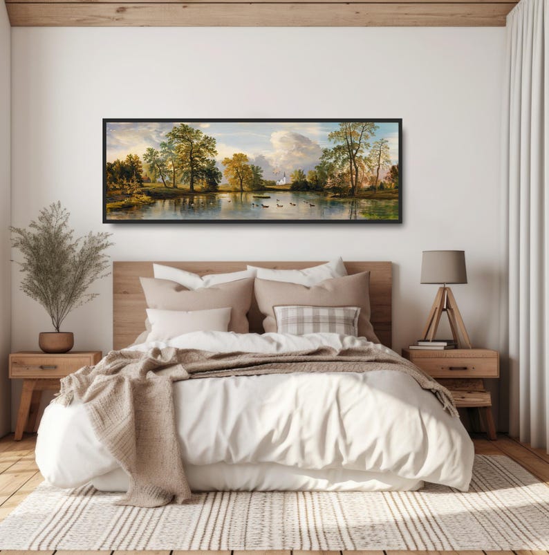 K&ouml;nnte beinhalten: Ein gerahmtes Landschaftsbild mit einem See, B&auml;umen und einem Geb&auml;ude in der Ferne. Das Bild h&auml;ngt an einer wei&szlig;en Wand &uuml;ber einem Bett mit wei&szlig;en Bettw&auml;sche und einer braunen Decke.