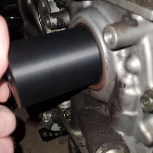 Op de afbeelding: Een close-up van een motoronderdeel. Een zwart cilindrisch onderdeel wordt in een metalen behuizing geplaatst. De motor is zilverkleurig met zichtbare olieresten.