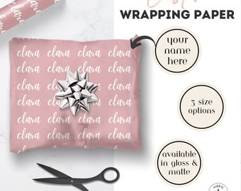 May include: Custom wrapping paper in a dusty pink shade featuring the repeating white name Clara in a cursive script. The set includes a roll of paper, a silver gift bow, and black scissors. Text on the image reads Custom WRAPPING PAPER.