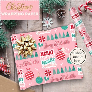Puede incluir: Papel de regalo de Navidad rosa con un patrón repetido de bastones de caramelo, árboles de Navidad, adornos y las palabras "Merry Merry Christmas". El papel está personalizado con el nombre "Lucy Michelle".