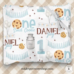 May include: A white baby blanket with a blue and brown pattern. The blanket features a milk jug, chocolate chip cookies, and the words "one sweet cookie" and "Happy Birthday Daniel Jay".
