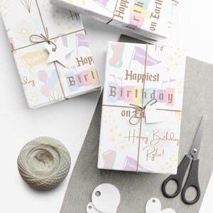 May include: Gift-wrapped presents with birthday-themed wrapping paper. The paper features pastel colors, flags, balloons, and the text "Happiest Birthday on Earth" and "Happy Birthday Rylie!". Presents are tied with twine and star-shaped tags.