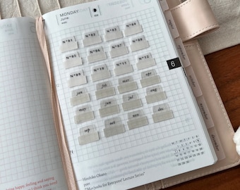 May include: A close-up of a planner with a grid pattern and clear plastic tabs labeled with numbers and months of the year. The planner is open to June and the tabs are labeled with numbers 1 through 12 and the months January through December. The planner is on a light pink surface.