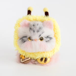 May include: A plush toy cat with a bee costume. The cat has gray and white fur, pink cheeks, and large, golden eyes. It wears a yellow fuzzy bee costume with antennae and a honey pot charm. The toy is on a white background.