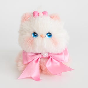 Może przedstawiać: Pluszowy biało-różowy kotek z dużymi niebieskimi oczami i różową kokardą. Kotek ma różowe akcenty na uszach i policzkach oraz małą różową kokardkę na głowie. Duża różowa satynowa kokarda zdobi jego szyję.