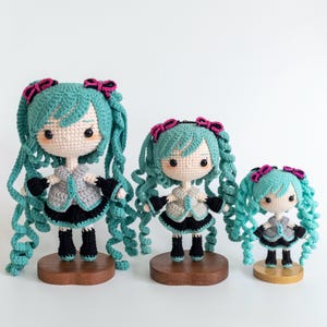 May include: Three crocheted dolls with teal hair, pink bows, and grey and black outfits stand on wooden bases. The dolls vary in size, with the largest doll measuring approximately 25 cm tall. The dolls have detailed facial features and are handmade.