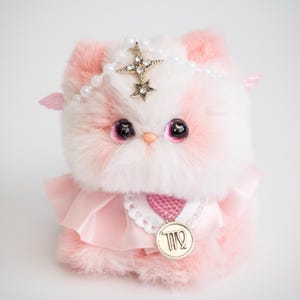 May include: A plush, pink and white cat doll with large, dark pink eyes. The doll wears a pearl and star crown, pink wings, and a pink dress with a Virgo pendant. The doll is made of soft, fluffy material.