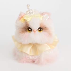 May include: A plush cat figurine with a crown, yellow bow, and collar. The cat has brown and white fur, large golden eyes, and pink cheeks. The collar is yellow with a lace trim. The crown is silver with a pearl on top.