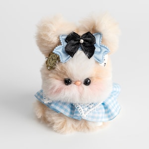 Może przedstawiać: Pluszowy królik z jasnobrązowym i białym futrem. Nosi niebieską sukienkę w kratkę z białym koronkowym kołnierzem, czarną kokardę z perłą i niebieską kokardę. Do głowy przymocowany jest mały urok zegara.