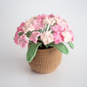 Puede incluir: Un arreglo de flores de ganchillo en una maceta marrón. Las flores son rosas y blancas con un centro de perla. Las hojas son verdes.