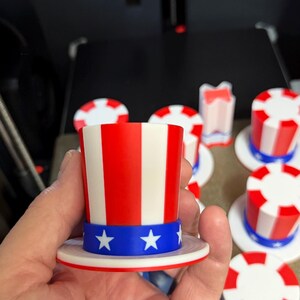 May include: A miniature Uncle Sam hat with red and white stripes and a blue band with white stars. The hat is held in a hand, with several similar hats in the background. The hats are likely for a patriotic theme.